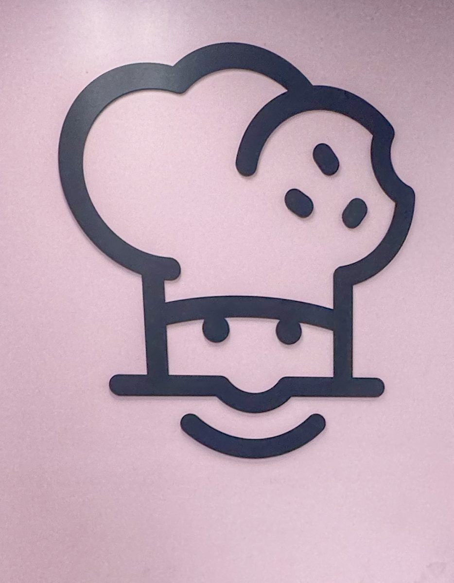 The Crumbl logo is a baker with a cookie in their hat. The large logo is visible to customers on the left side of the wall near the order pickup area.